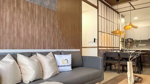 A modern living room with a grey sofa, a white cushion with a logo, and a wooden accent wall. - Ancestry 3BR @ M Vertica | MRT Access+Netflix+WiFi (Cheras)