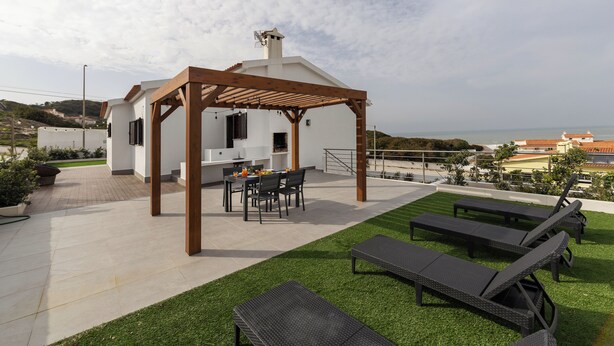 A modern outdoor patio with a pergola, a dining area, and lounge chairs. - Lagoon View Villa - Ocean Serenity (Óbidos)