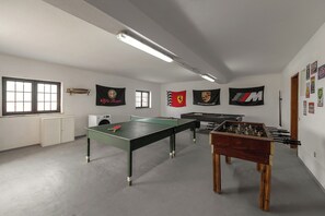 Una habitació amb taula de ping-pong, taula de futbolí i taula de billar. Hi ha banderes a les parets i una finestra amb persianes.