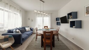 A modern living room with a blue sofa, a dining table with chairs, and a large window with sheer curtains. - Lagoon View Villa - Ocean Serenity (Óbidos)