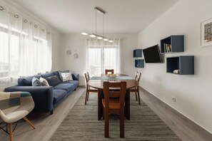 Un saló modern amb un sofà blau, una taula de dinar amb cadires i una finestra gran amb cortines lleugeres.