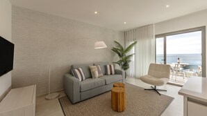 Un salon moderne avec un canapé gris, une table d’appoint en bois et une grande fenêtre donnant sur la mer.
