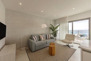 A modern living room with a grey sofa, a wooden side table, and a large window with a view of the sea.