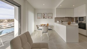 Une cuisine moderne avec des meubles blancs, un coin repas doté d’une table ronde et de chaises, et une vue sur un paysage côtier grâce à de grandes fenêtres.