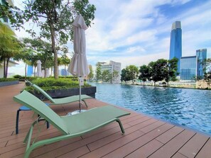 Zona junto a la piscina con sillones de descanso y una sombrilla, rodeada de vegetación y edificios modernos al fondo.