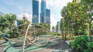 Ein Spielplatz mit einer Kletterkonstruktion aus Seilen, umgeben von Grünflächen und modernen Gebäuden im Hintergrund.