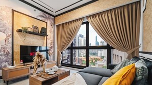 Una sala de estar moderna con un televisor grande, un sofá con cojines, una mesa de centro y vistas de la ciudad a través de grandes ventanales.