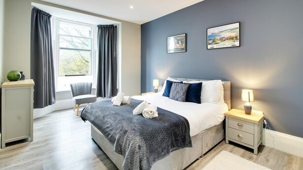 A modern bedroom with a large bed, bedside tables, a chair, and a window with curtains. - Regent Parade Retreat, sleeps 6, Free parking, (North Yorkshire)