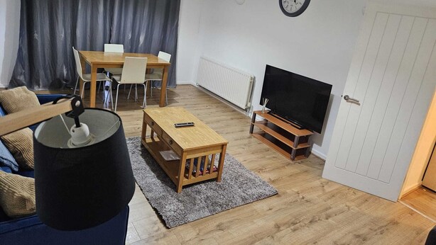 A living room with a wooden dining table, chairs, a TV on a stand, a sofa, a coffee table, and a large window with curtains. - Modern 2-Bed Apt Near Hospital and University of Birmingham (Birmingham)