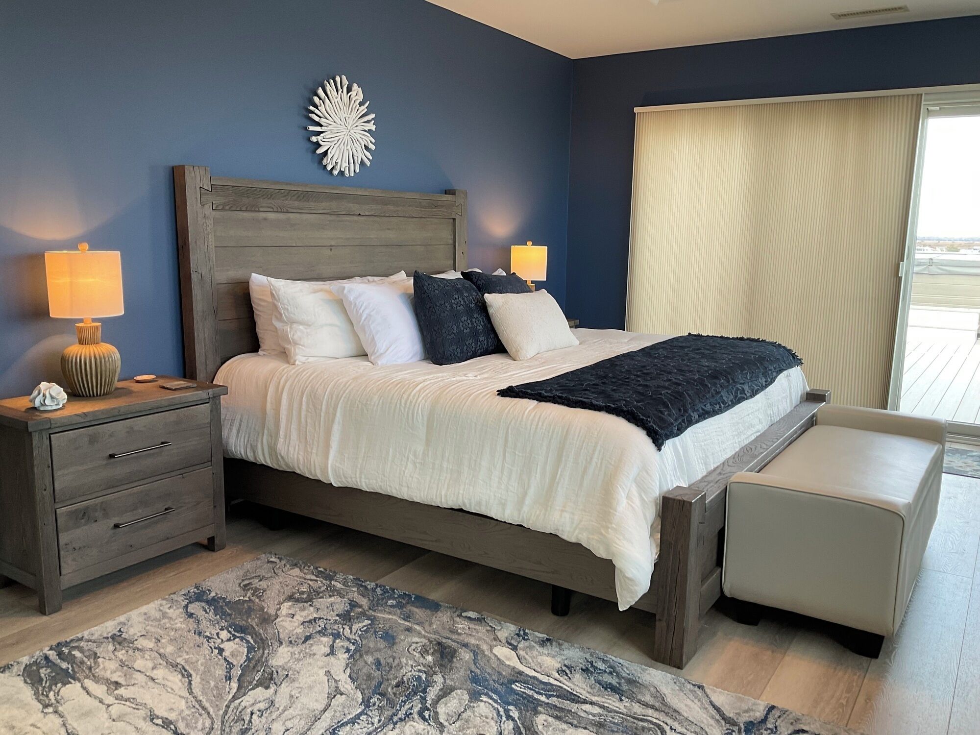 Un dormitorio con cabecera de madera, una cama grande con almohadas blancas y oscuras, una mesita de noche con una lámpara, un banco al pie de la cama y vistas al exterior a través de una ventana.