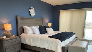 A bedroom with a wooden headboard, a large bed with white and dark pillows, a bedside table with a lamp, a bench at the foot of the bed, and a view of the outdoors through a window. - Penthouse Apartment, Ocean View & Pool View, 4 BR, 3 Bath (Wildwood Crest)