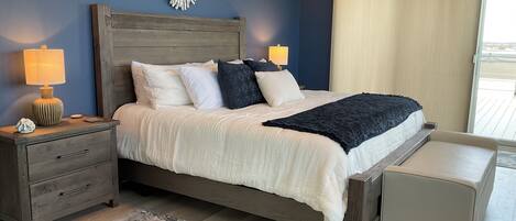 Un dormitorio con cabecera de madera, una cama grande con almohadas blancas y oscuras, una mesita de noche con una lámpara, un banco al pie de la cama y vistas al exterior a través de una ventana.