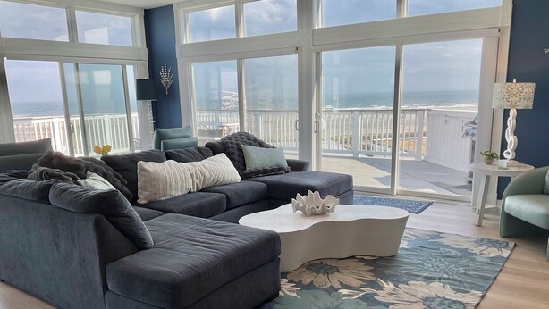 A spacious living room with a dark grey sectional sofa, a white coffee table, and a large window overlooking a balcony with a view of the ocean. - Penthouse Apartment, Ocean View & Pool View, 4 BR, 3 Bath (Wildwood Crest)