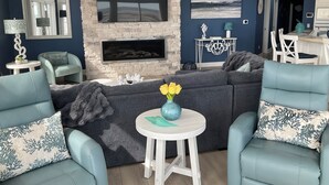 A living room with a stone fireplace, a large flat-screen TV, and a dining area in the background. - Penthouse Apartment, Ocean View & Pool View, 4 BR, 3 Bath (Wildwood Crest)