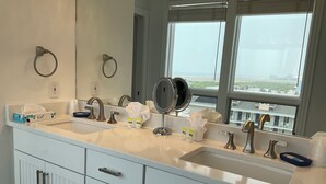 A bathroom with a double sink, two towel rings, a mirror, and a window with a view of buildings. - Penthouse Apartment, Ocean View & Pool View, 4 BR, 3 Bath (Wildwood Crest)
