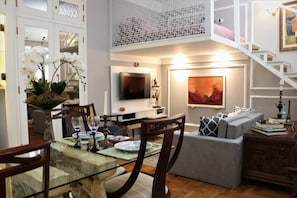 Ein modernes Wohnzimmer mit einem Glastisch, Holzstühlen, einem grauen Sofa, einem Fernseher und einer Treppe.