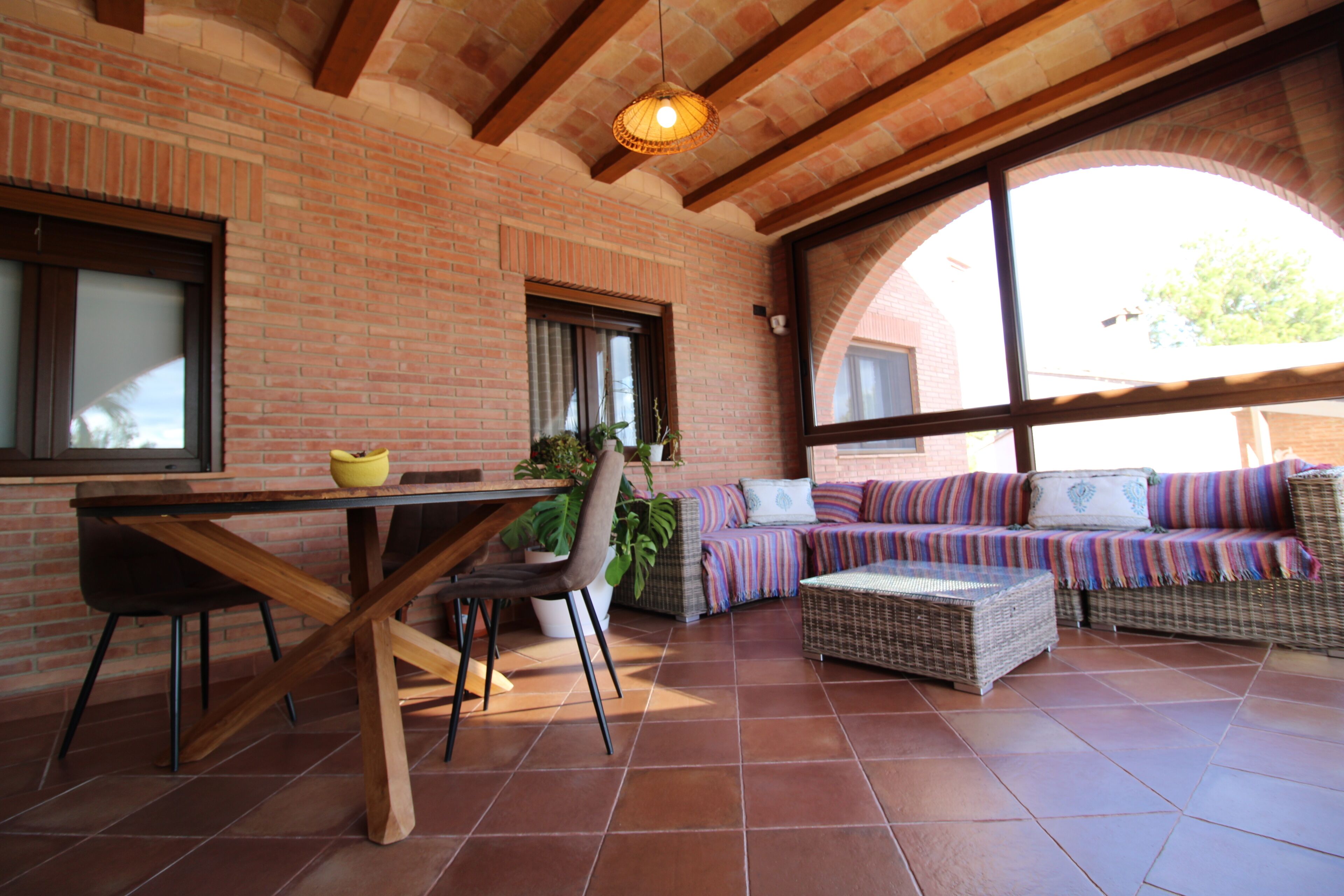 Een terras met een bakstenen muur, houten balken, een tafel met stoelen, een bank en rieten meubels.