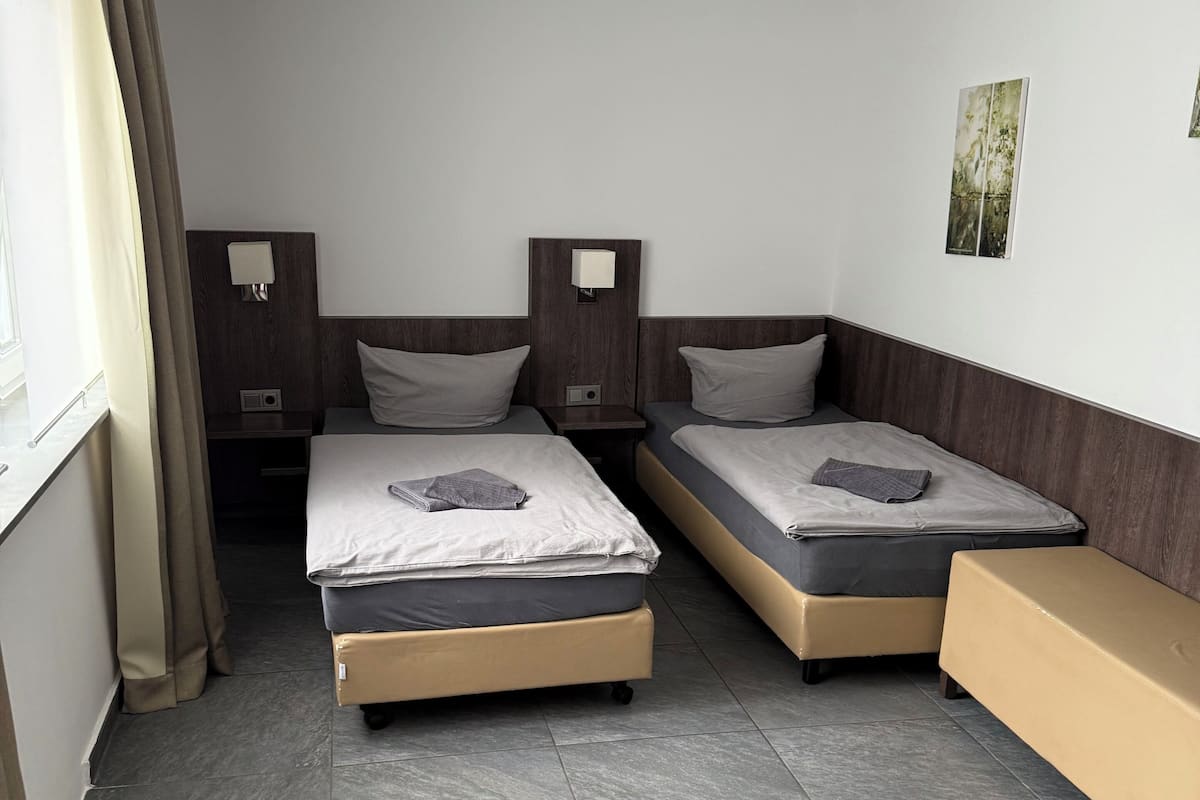 Una camera d'albergo con due letti, una panca, una finestra con tende e un quadro appeso al muro.