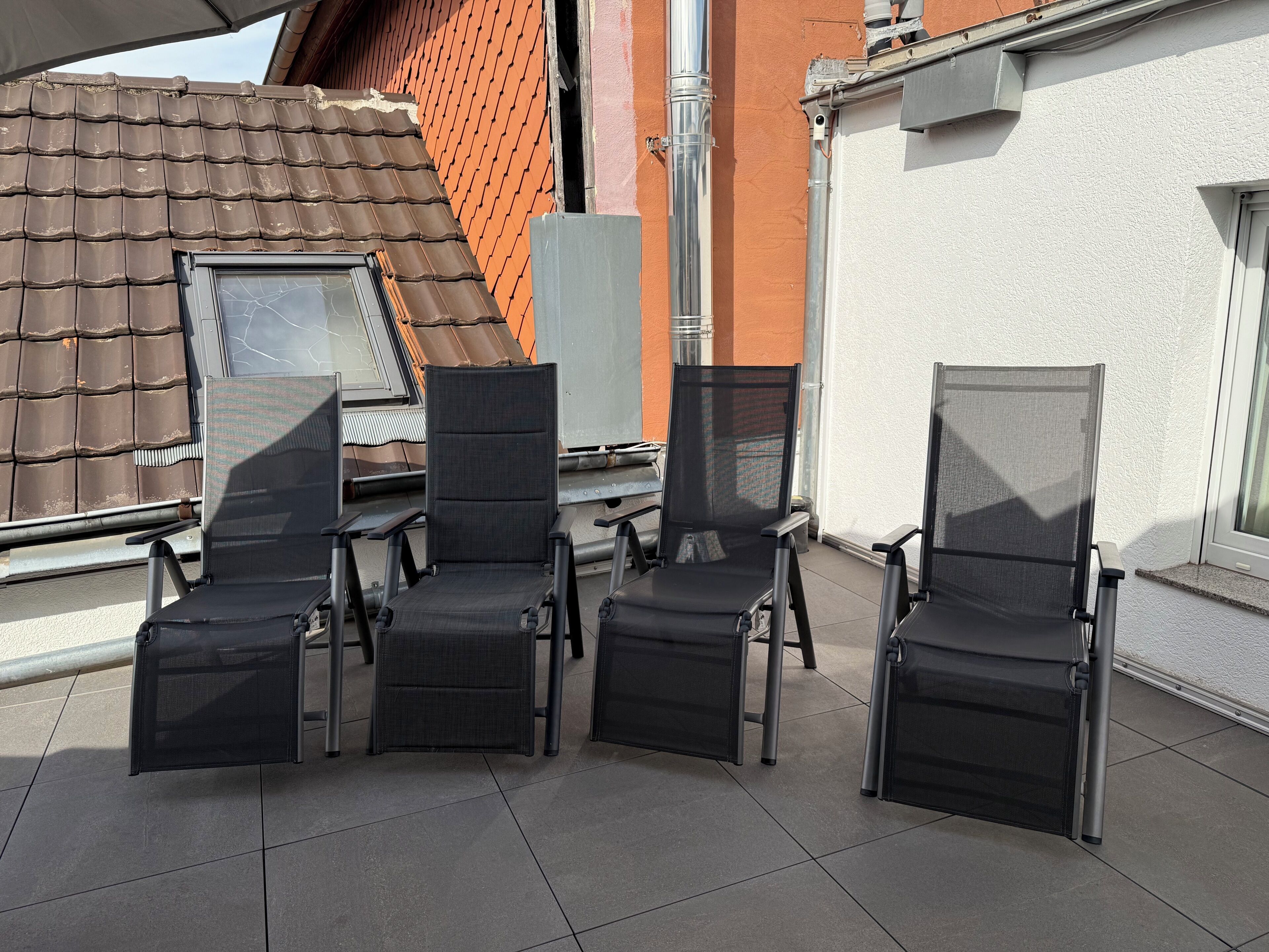 Una terraza en la azotea con cuatro sillones de descanso negros, una mesa y un ventanal con vistas a un tejado de tejas rojas.