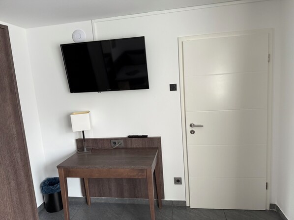 A room with a flat-screen TV mounted on the wall, a wooden table with a lamp, a door, and a trash bin. - Blauer Stein - Core it02- Haus (Bergheim)