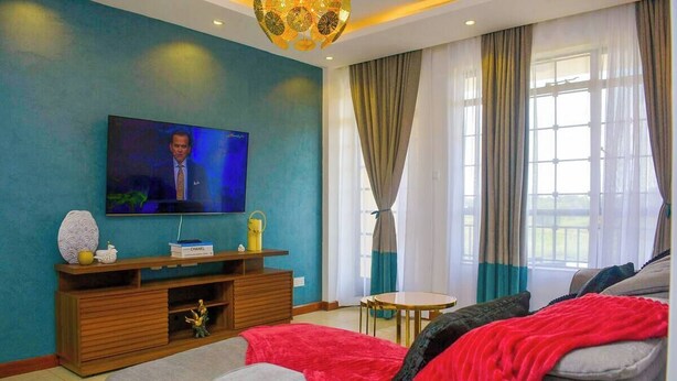 A modern living room with a flat-screen TV, wooden entertainment unit, grey sofa, red throw blanket, and large windows with curtains. - 'THE TEAL' PREMIUM FURNISHED LUXURY 2-BEDROOM APARTMENT — MOVE-IN READY (Kenya)