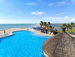A pool with a curved edge, surrounded by lounge chairs and palm trees, overlooking a beach and ocean. - Ocean Breeze Home for 16 guests! (Laguna Shores)