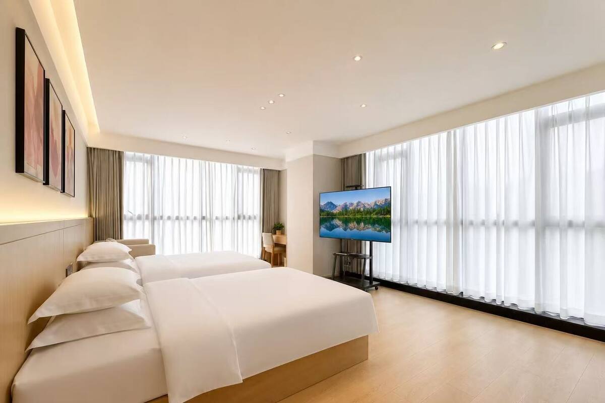 A modern hotel room with a large bed, a flat-screen TV, and large windows with sheer curtains.