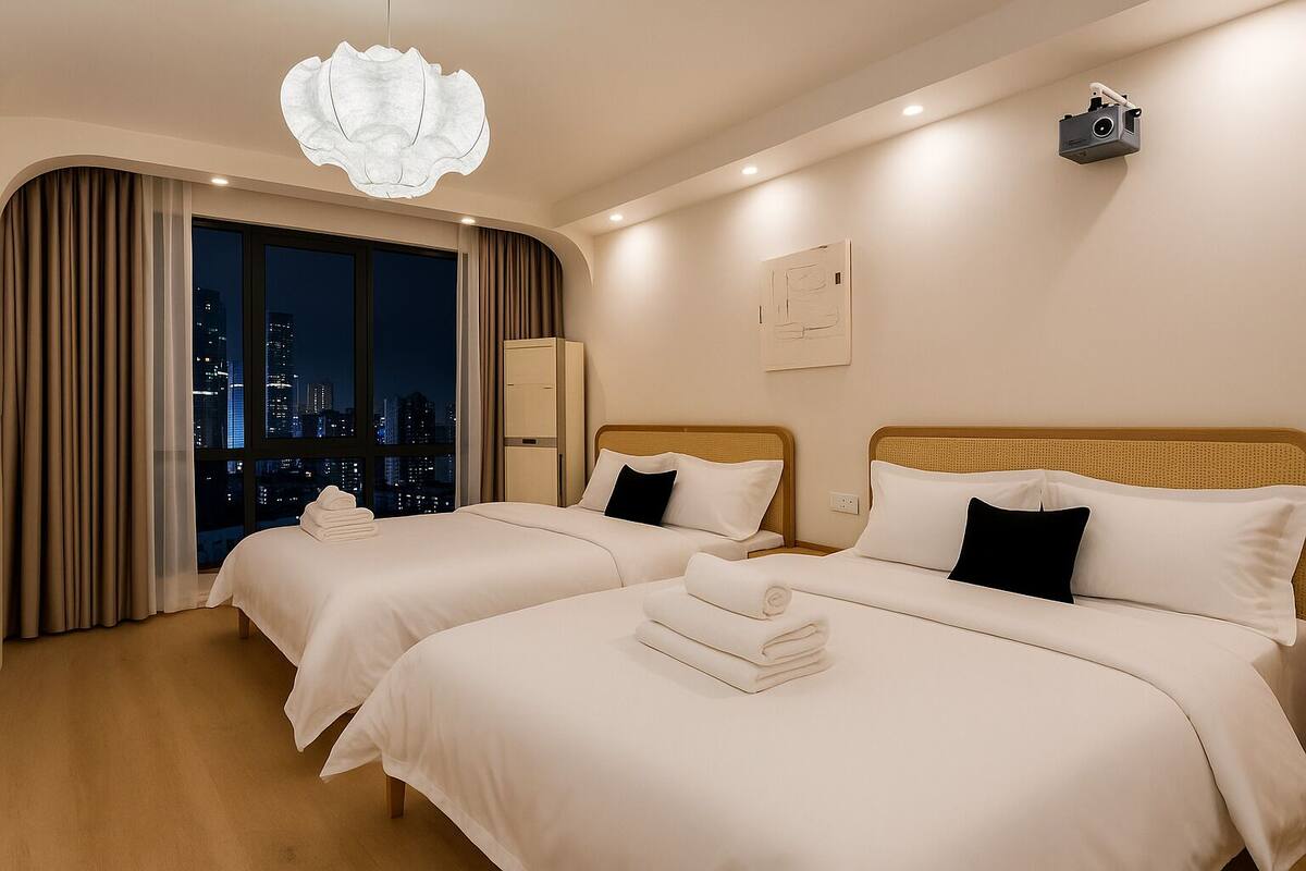 A hotel room with two beds, a city view, a large window, and a projector on the wall.