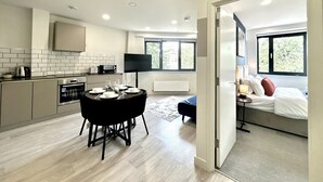 A modern kitchen with a dining area, a large window, and a bedroom with a bed and window seat. - Central 2 Bed Flat, Free Parking, Gym, Cinema Room (Bedford)