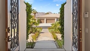 A gated driveway leading to a house with a garage, surrounded by a landscaped garden with palm trees and flowering plants. - Desert Luxe Oasis: Private Pool and 4+ BR in Gated South La Quinta! (La Quinta)