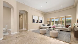 A spacious living room with a beige sofa set, ottomans, and a coffee table. There are two large windows with blinds, a fireplace, and a hallway leading to another room. - Desert Luxe Oasis: Private Pool and 4+ BR in Gated South La Quinta! (La Quinta)