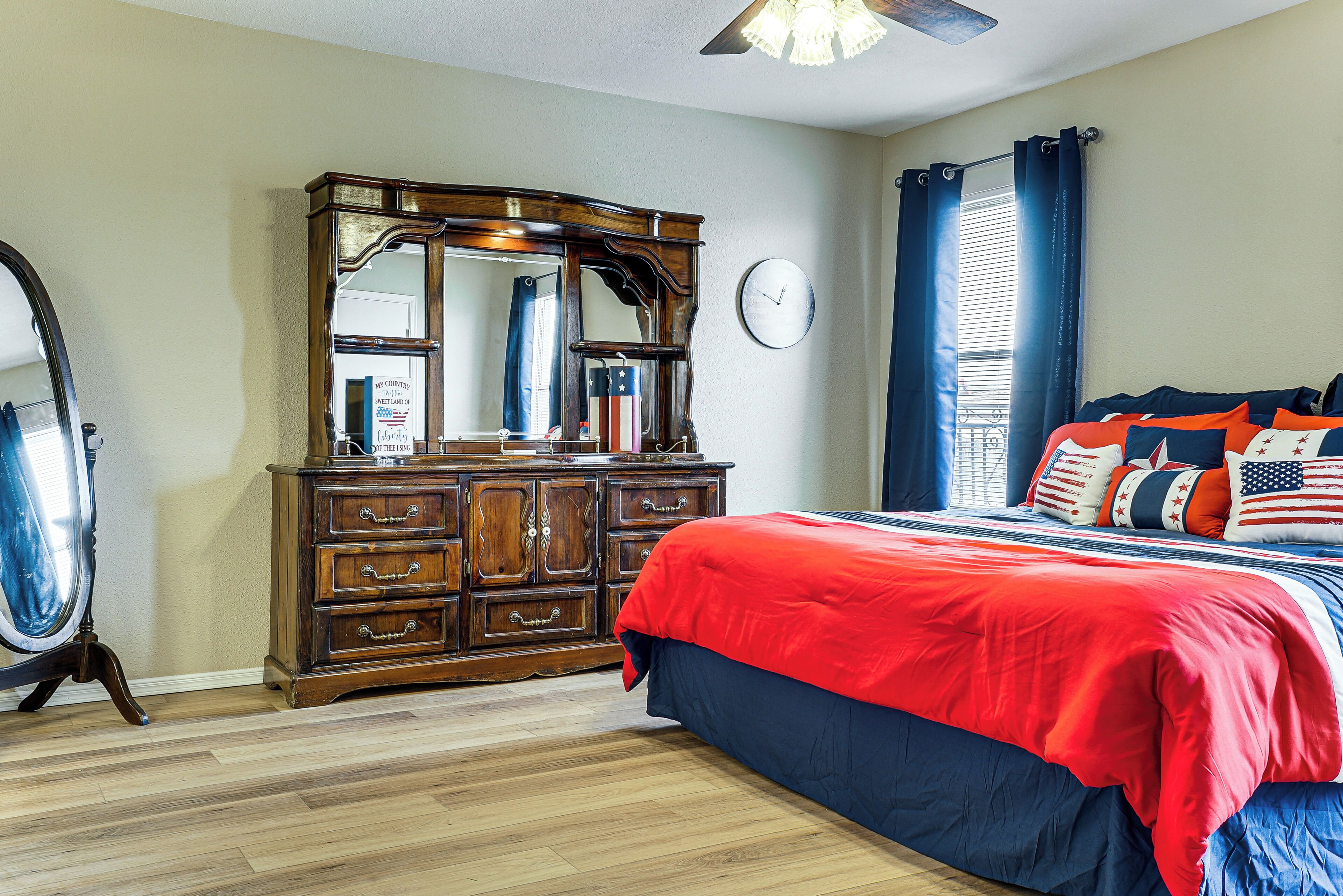 A bedroom with a wooden dresser, a bed with red and blue bedding, a mirror, and a clock on the wall.