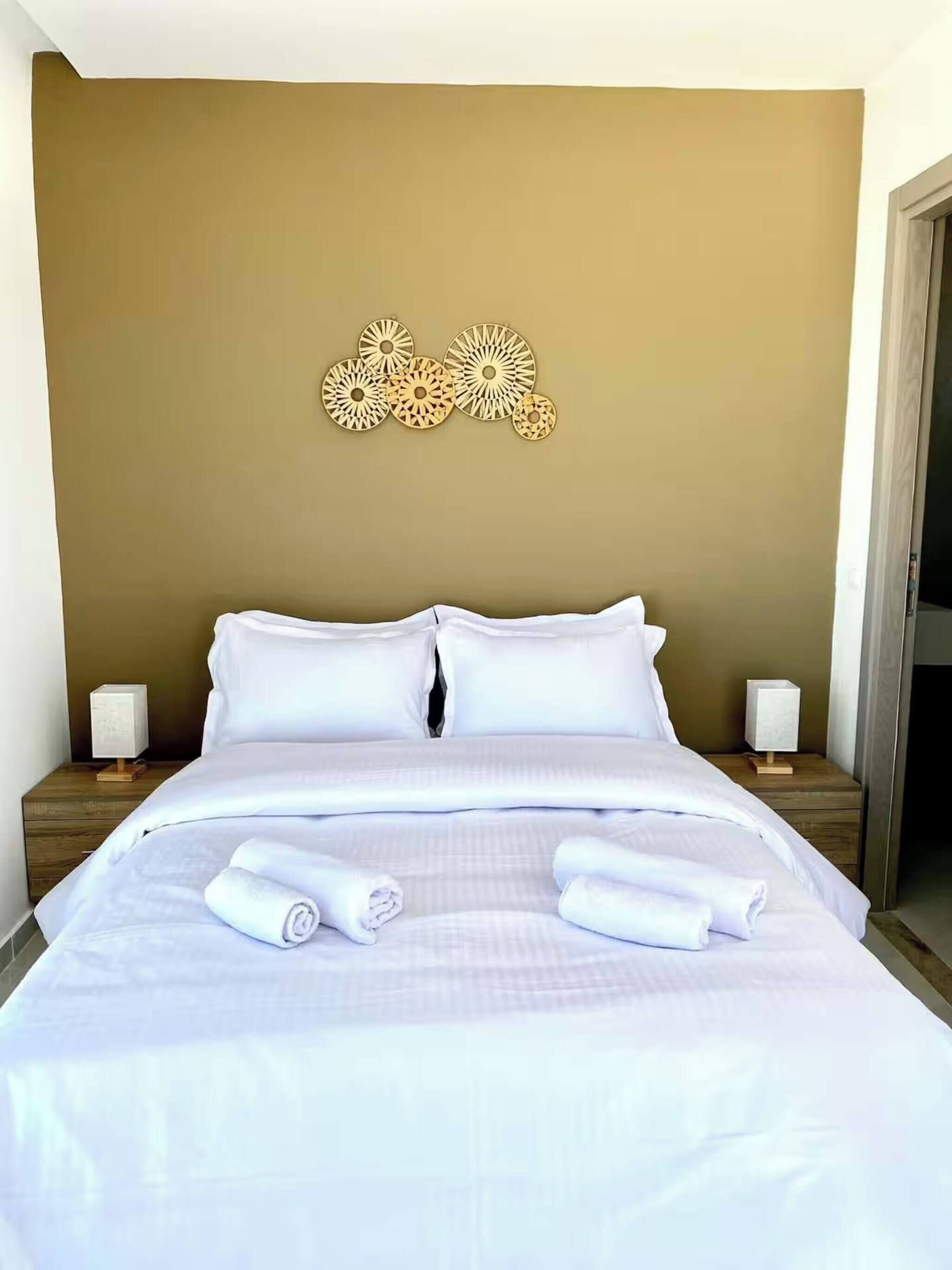 Un lit bien fait, avec du linge de lit blanc et des serviettes pliées, un mur brun orné de décorations murales et deux tables de chevet avec des lampes.