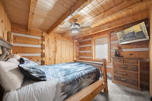 Quarto de cabana de toras com uma cama, cômoda, ventilador de teto e janela.