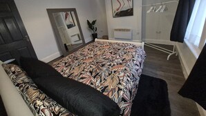 Et soveværelse med seng, et spejl, en potteplante, et vindue med gardiner og en fastgjort varmeovn.