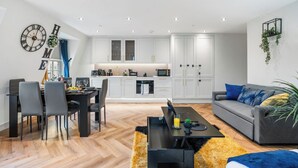 A modern living room with a sofa, dining table, and chairs, a kitchen with white cabinets, and a clock on the wall.