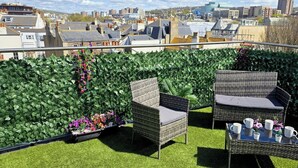 A rooftop terrace with green foliage, wicker furniture, and a table set with cups and flowers.