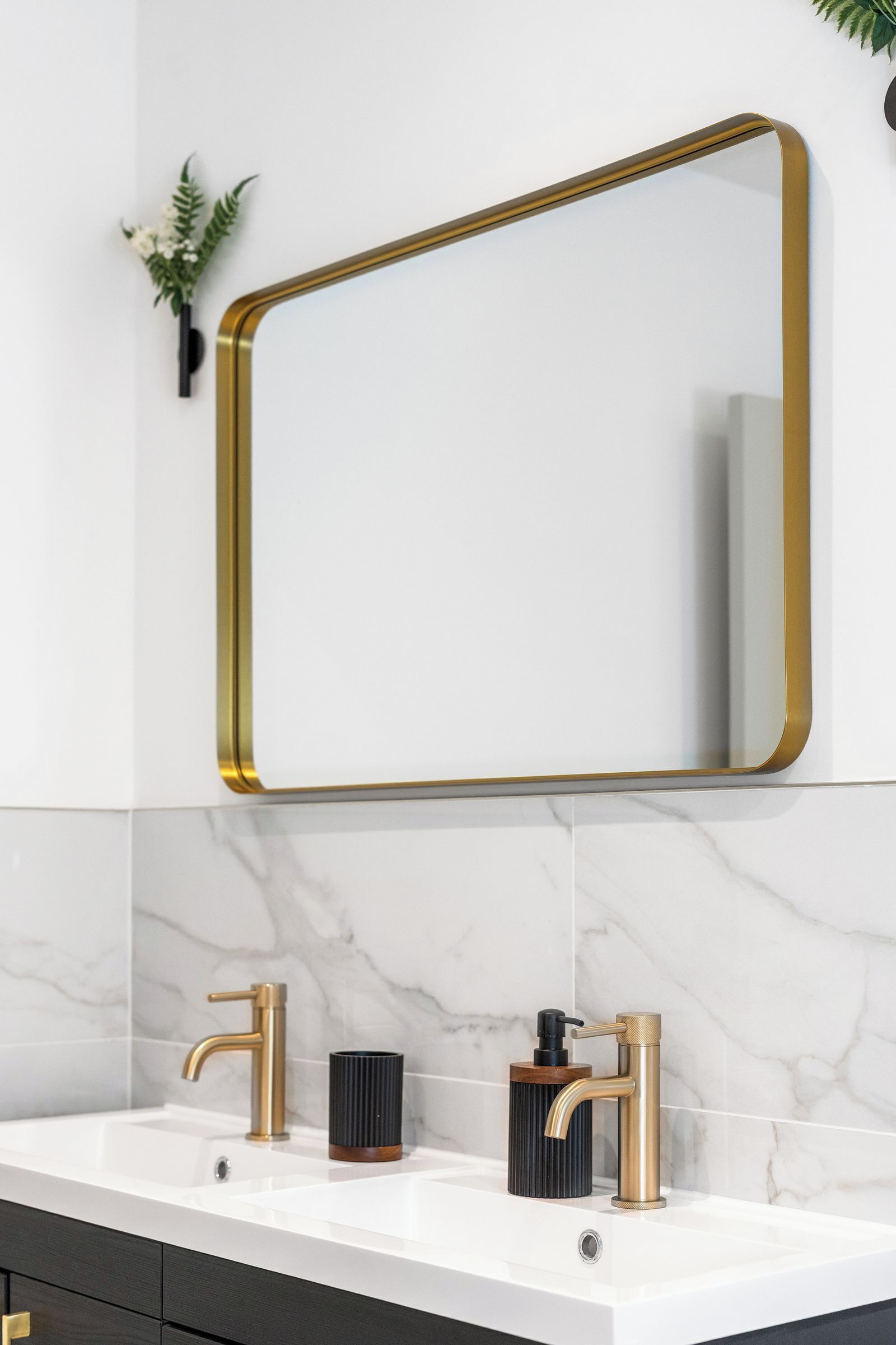 A bathroom with a white marble sink, two gold faucets, a black soap dispenser, and a gold-framed mirror.