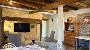A rustic interior with exposed stone walls, a wooden ceiling, a flat-screen TV mounted on the wall, and a dining area with a table and chairs. - Gîtes de France® - Héloïse's gîte (BETAILLE)