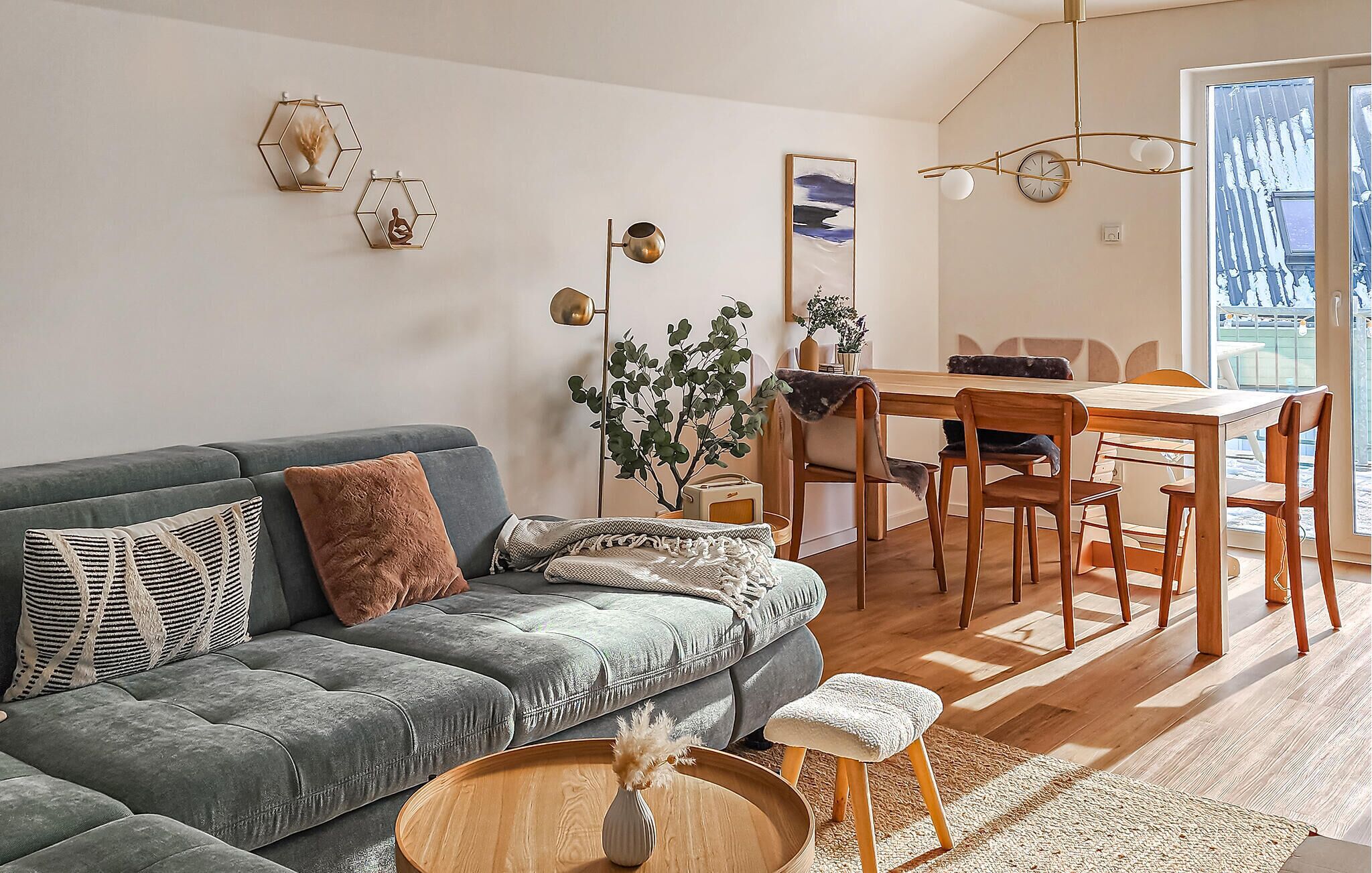 En moderne stue med en grå sofa, et træspisebord og stole. Der er en plante, en vase og en hængende lampe.