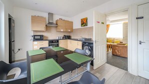 Een praktische keuken met een magnetron, kookplaat en wasmachine, een eethoek met groene placematen, en uitzicht op een woonkamer met een bank en gele kussens.