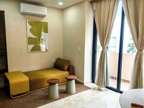 Ein modernes Zimmer mit einer Couch, zwei Hocker, einem großen Fenster mit Vorhängen, einer Klimaanlage und einem an der Wand hängenden Kunstwerk.