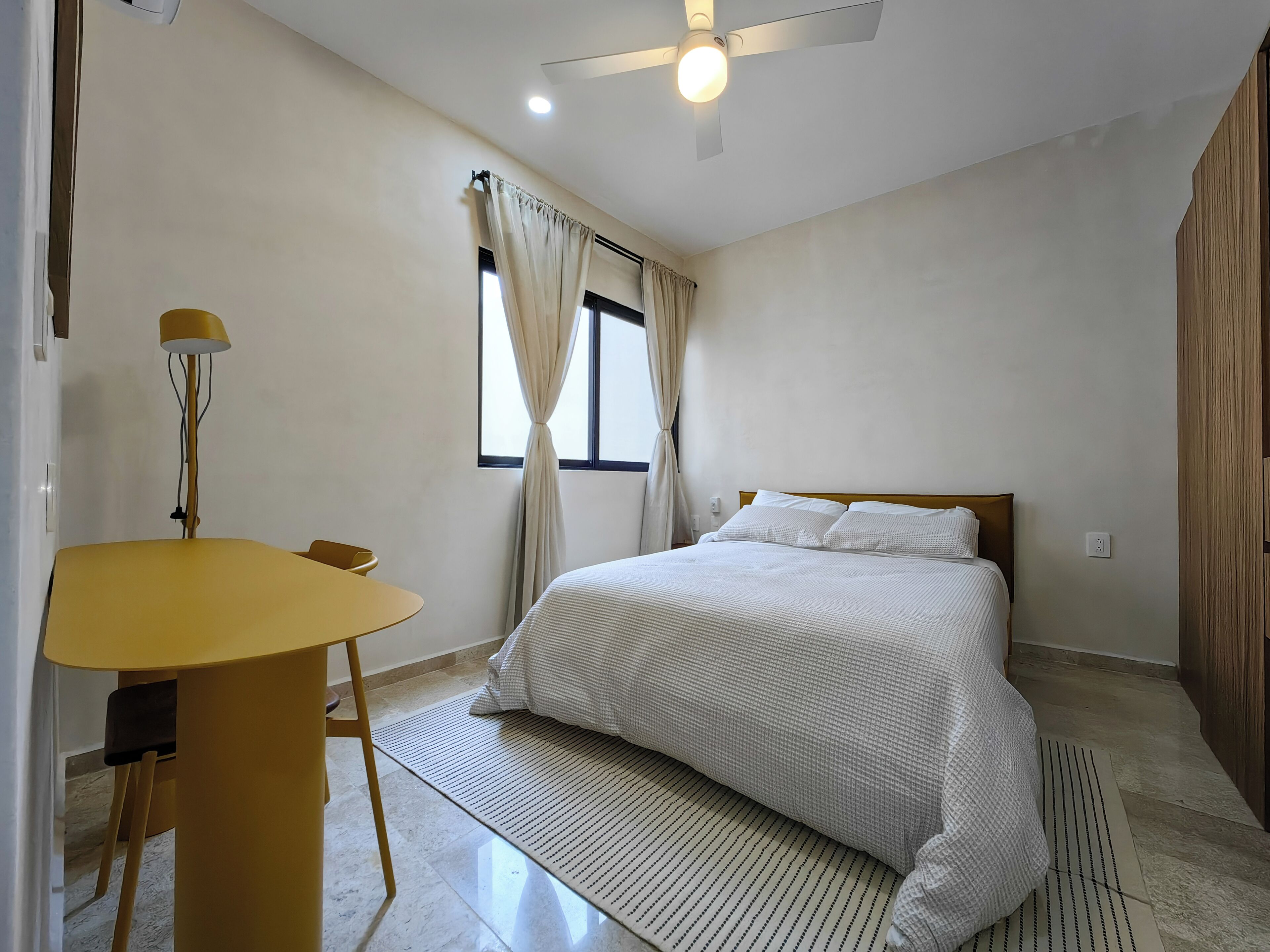 Ein Schlafzimmer mit einem Bett, einem Schreibtisch, einem Stuhl, einem Deckenventilator und einem Fenster mit Vorhängen.