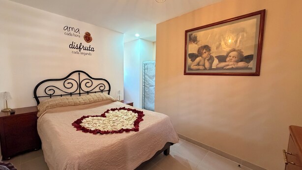 Un dormitorio con cama, mesita de noche, lámpara, un cuadro en la pared y un corazón decorativo hecho de pétalos de rosa sobre la cama.