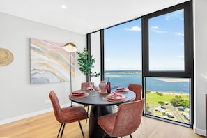 A dining area with a round table, four chairs, a large painting, a floor lamp, and a view of the sea through large windows. - Broadwater & Ocean Views 2BR Apartment Southport (Southport)