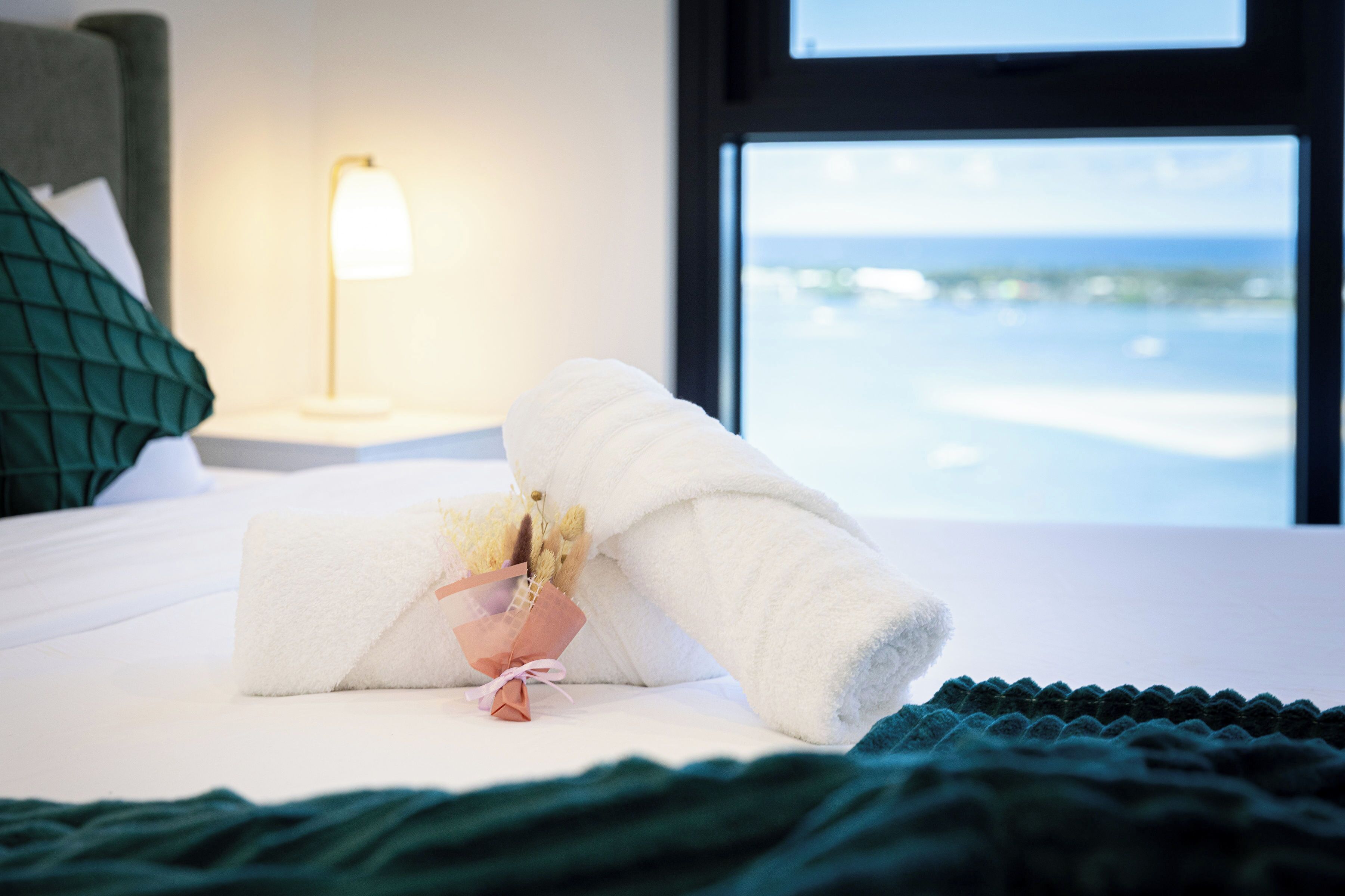 A neatly made bed with a towel and a small bouquet of flowers, a green pillow, a bedside lamp, and a view of the sea through a window.