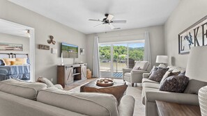 A spacious living room with a large sofa, a coffee table, and a television mounted on the wall. There is a sliding glass door leading to an outdoor area with a view of trees and a building. - Lakeside Sunrise Water Views (Camdenton)