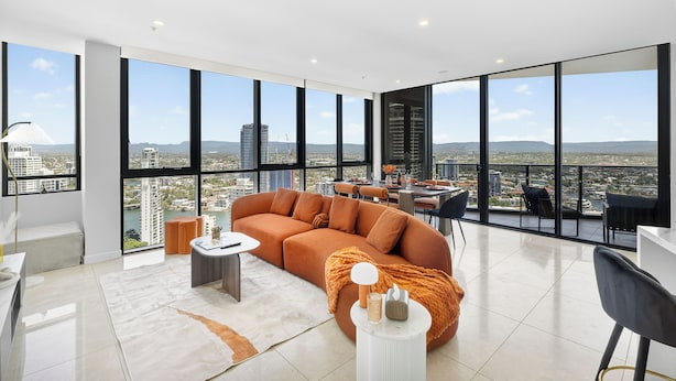 A modern living room with a large sofa, a dining area, and a view of the cityscape through large windows. - Ruby 30F Sunset Penthouse | Ocean & City Views (Surfers Paradise)
