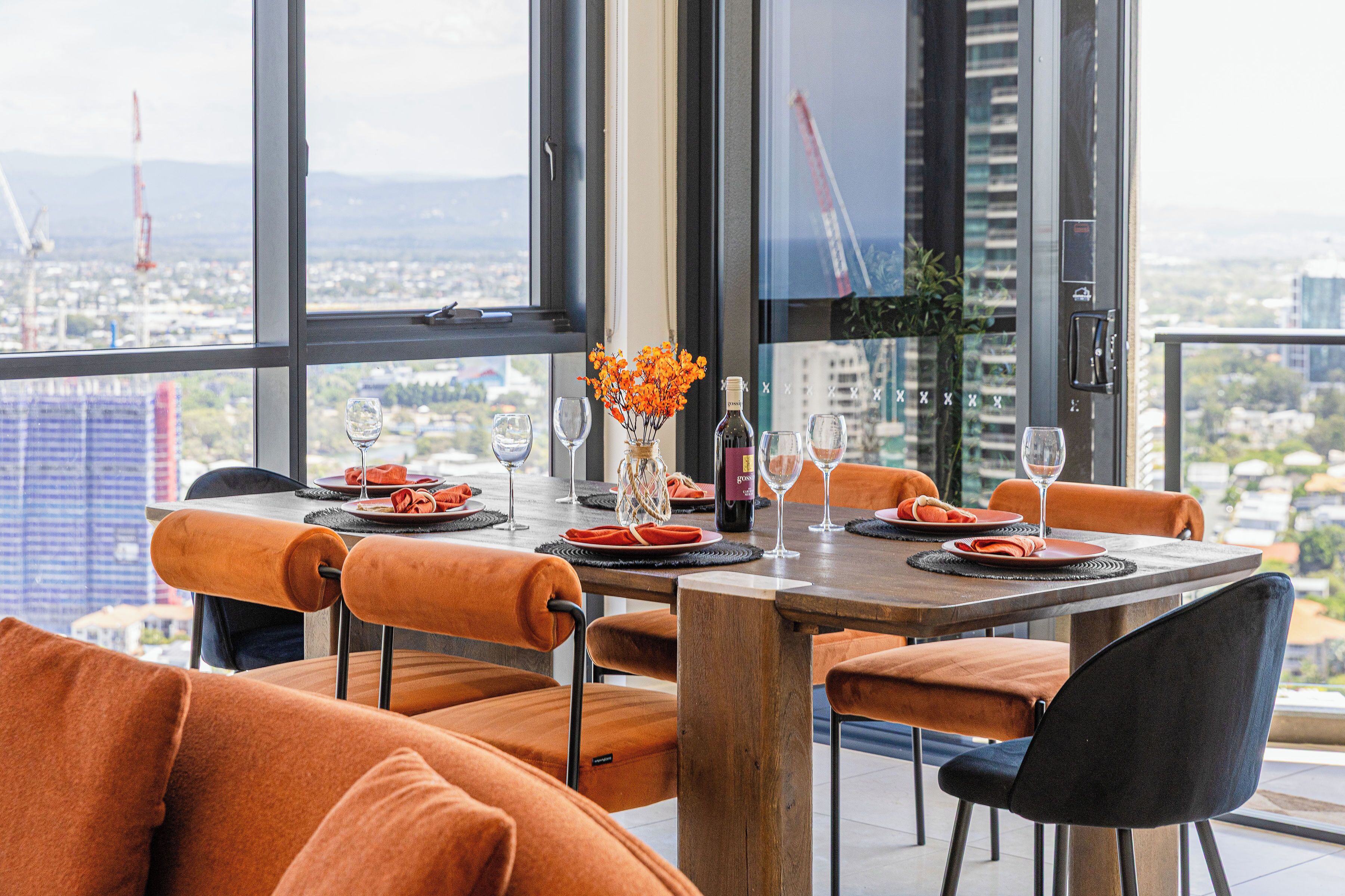 A dining area with a table set for four, large windows, orange upholstered chairs, and a view of the cityscape.