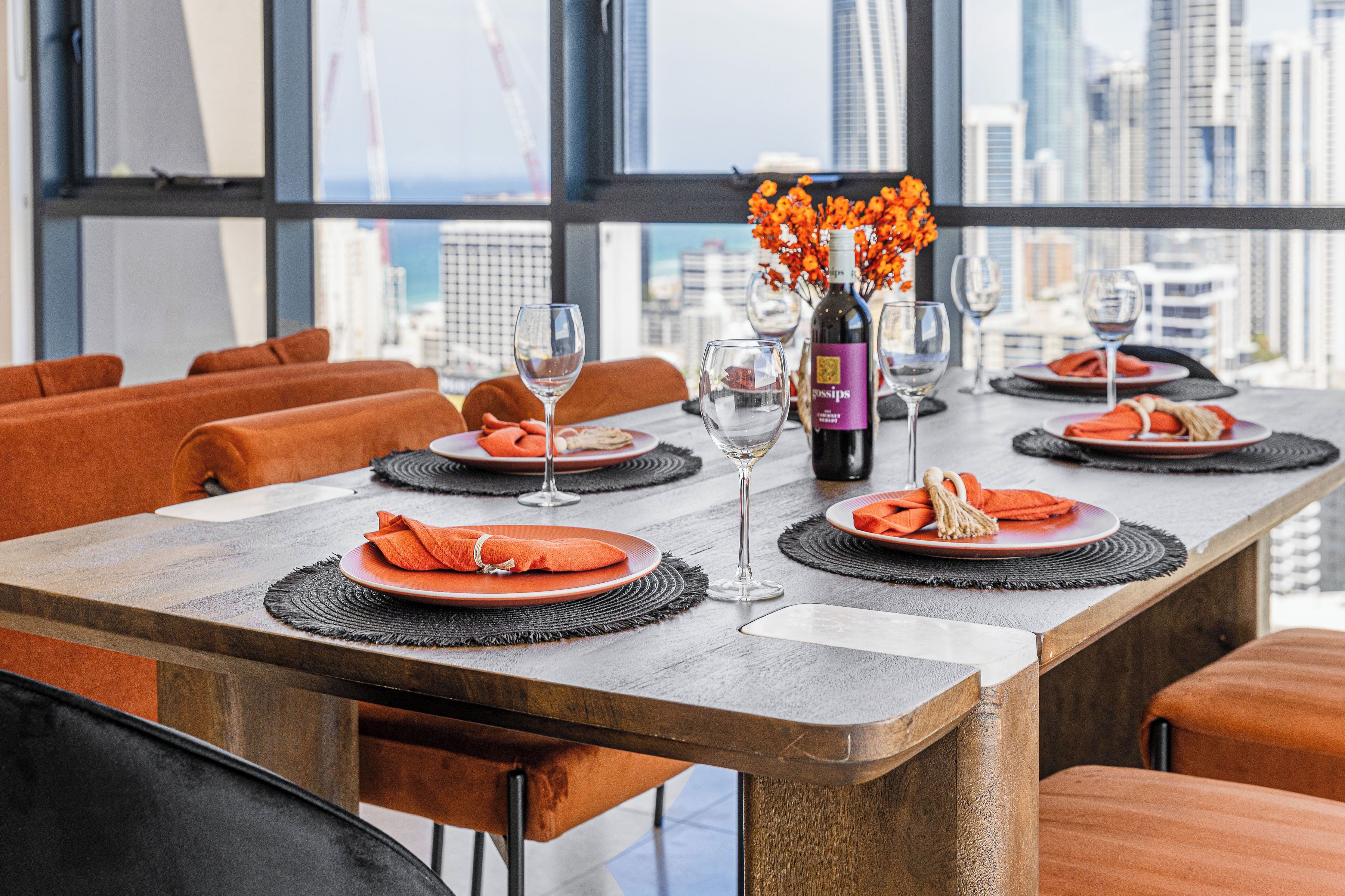 Un coin repas avec une longue table dressée pour six personnes, agrémenté de coussins orange, de sets de table noirs et d’une bouteille de vin.